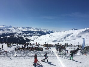 Pista de esqui coberta de neve com esquiadores e snowboarders, um teleférico e montanhas ao fundo.