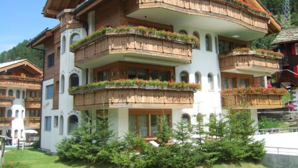 Ett hotell med flera våningar, vars balkonger är prydda med blommor, och som har en välskött trädgård och en imponerande bergskuliss i bakgrunden.
