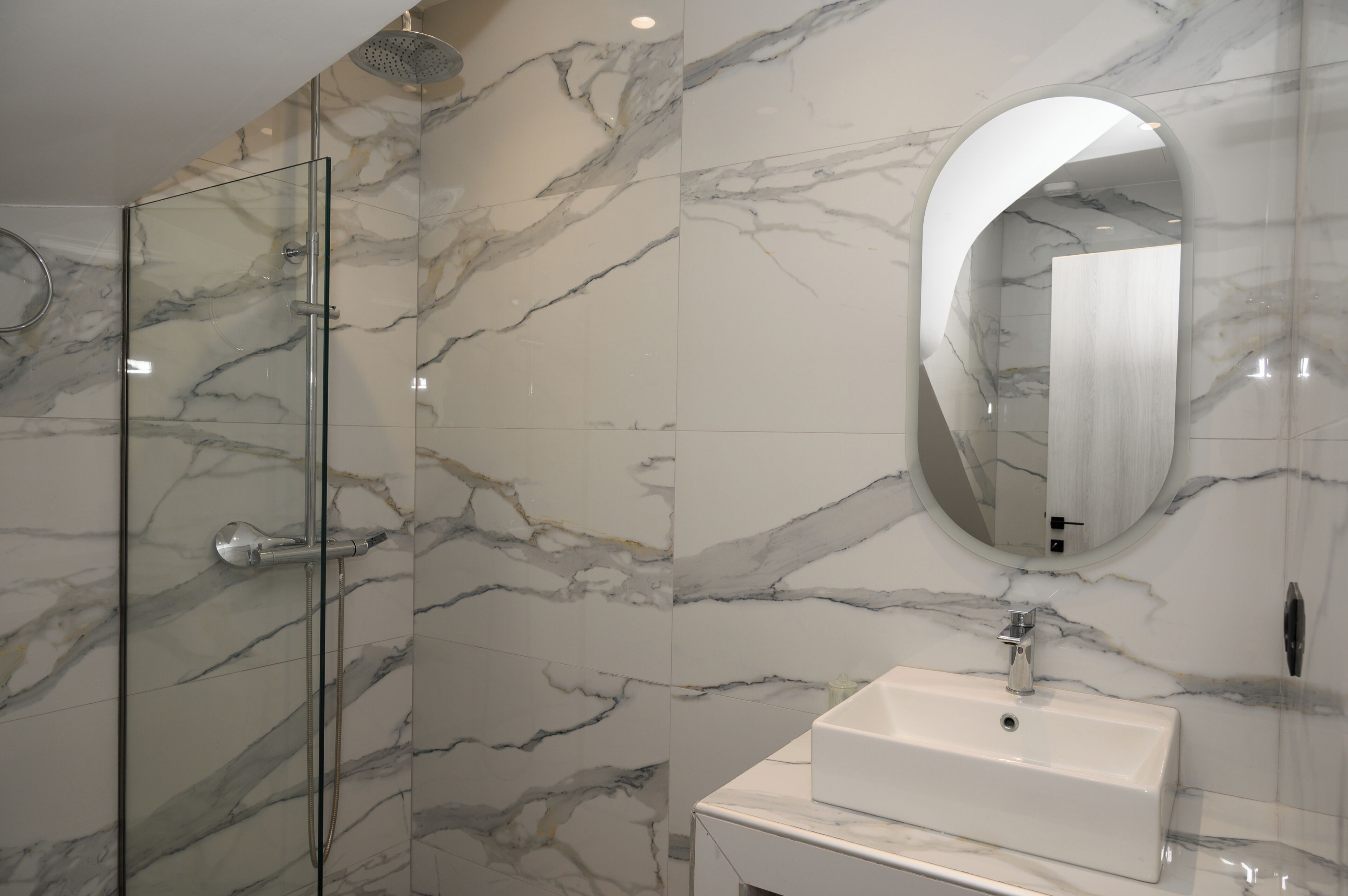 A modern bathroom with a glass shower enclosure, a marble-tiled wall, and a round mirror.