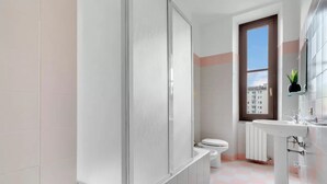 A bathroom with a shower, toilet, and sink, with a checkered floor and a window. - CO14 [Central] • Comfort • (Milano)