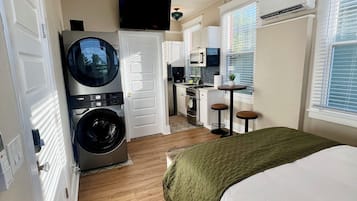A compact hotel room with a bed, a washing machine, a kitchenette, and a ceiling fan.
