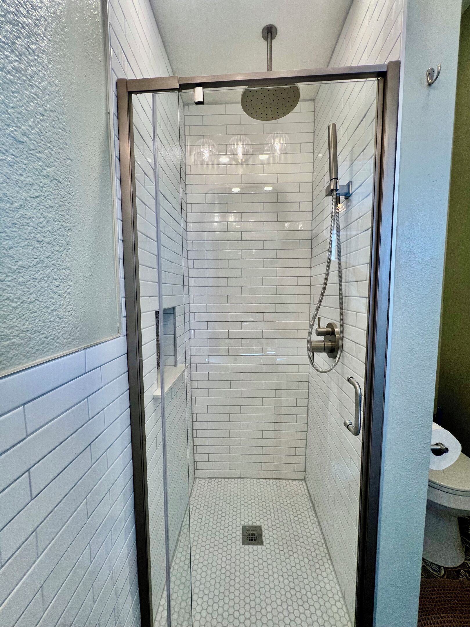 A glass-enclosed shower stall with white subway tiles, a handheld showerhead, and a floor drain.