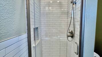 A glass-enclosed shower stall with white subway tiles, a handheld showerhead, and a floor drain.