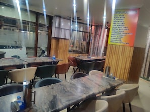 A restaurant interior with tables and chairs, a menu board listing various dishes, and a view of the street outside. - Hotel Maa Rudrani (Katra)