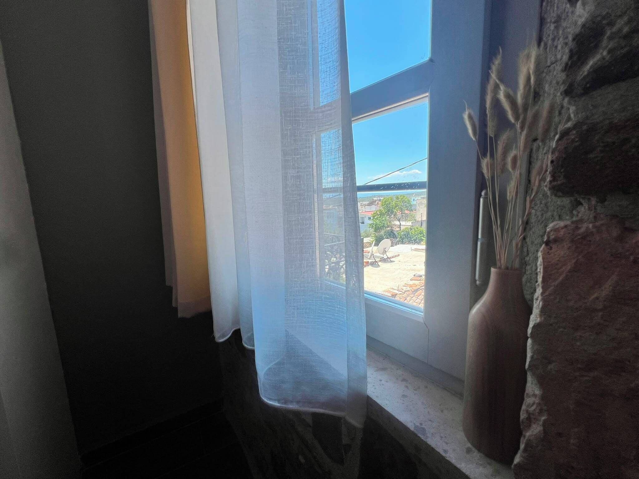 A window with sheer curtains, a stone wall, and a vase with dried plants.