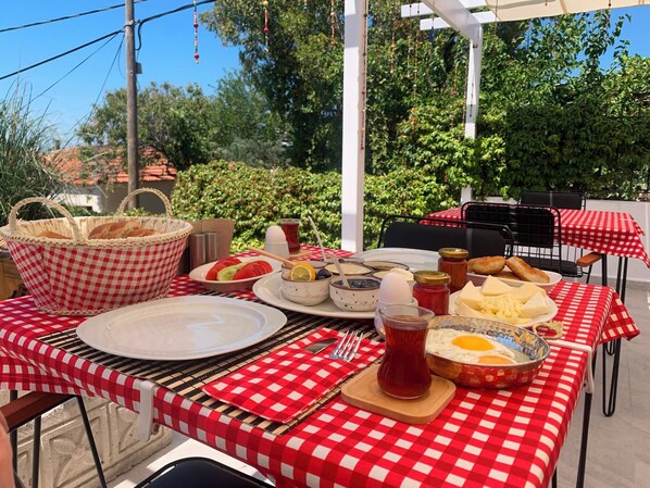 An outdoor dining area with red and white checkered tablecloths, a basket of bread, plates of food, and a variety of condiments.