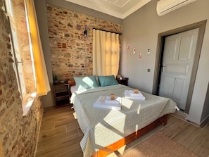 A bedroom with a stone wall, a bed with a patterned bedspread, a nightstand, a lamp, and a door.