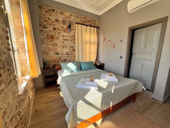 A bedroom with a stone wall, a bed with a patterned bedspread, a nightstand, a lamp, and a door.