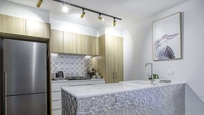 Eine moderne Küche mit einem Edelstahl-Kühlschrank, Holzschränken, einer weißen Kücheninsel mit gemustertem Spritzschutz und einem Bilderrahmen mit Vogelmotiv an der Wand.