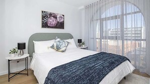Ein Schlafzimmer mit einem Bett, einem Nachttisch, einer Lampe, einem Fenster mit durchsichtigen Vorhängen und einem Bild an der Wand.