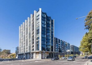 A modern multi-story building with a glass facade, located at a street corner with cars parked in front. - Great views - Apartment in Canberra CBD (Reid)