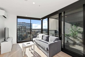 A modern living room with a grey sofa, a small white table, and a large window offering a city view. - Great views - Apartment in Canberra CBD (Reid)
