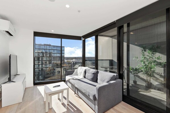 A modern living room with a grey sofa, a small white table, and a large window offering a city view. - Great views - Apartment in Canberra CBD (Reid)