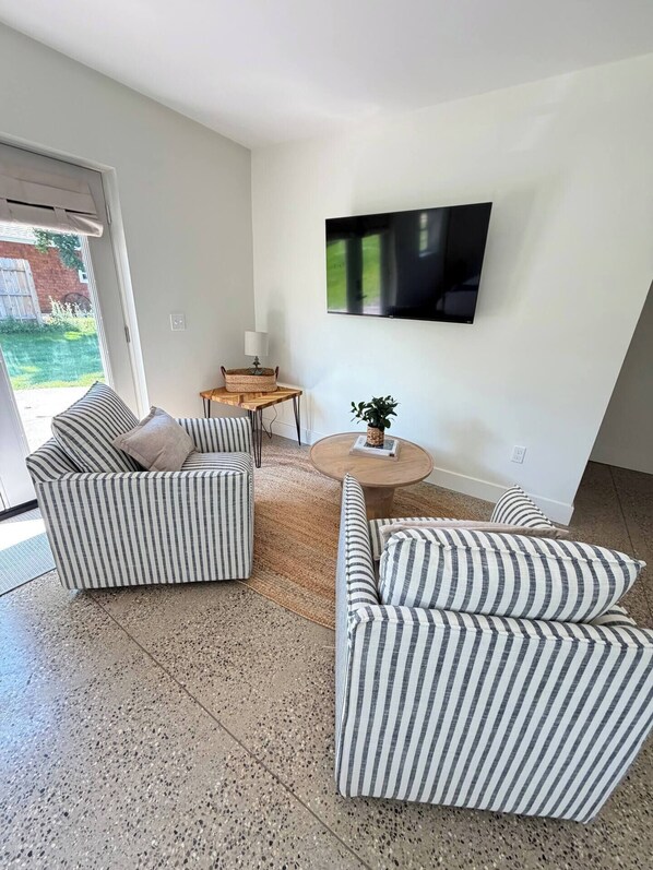A living room with striped sofas, a flat-screen TV mounted on the wall, and a small round table with a lamp.