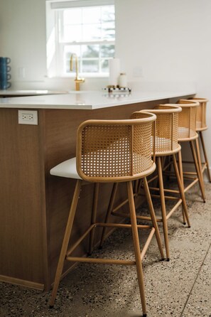 Un coin cuisine doté d'un plan de travail blanc, d'une fenêtre et de quatre tabourets de bar en osier avec des assises blanches.