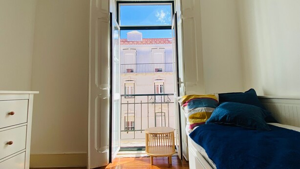 Un dormitorio con cama, una silla y vistas a edificios a través de una puerta abierta.