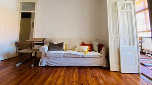 Una habitación con piso de madera, un sofá blanco con cojines, una pequeña mesa de madera y una puerta blanca entreabierta.