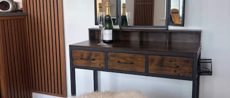 A wooden vanity with a mirror, a bottle of champagne, and a fluffy white pouf.