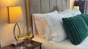 A neatly made bed with white linens and a green throw blanket, a bedside table with a lamp and two glasses of white wine, and a wooden headboard.
