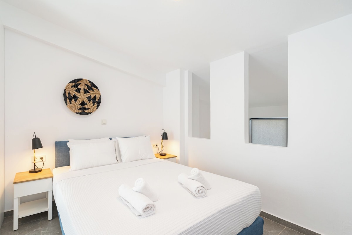 Quarto de hotel com cama, duas mesinhas de cabeceira, uma luminária fixada na parede, uma peça decorativa circular feita de fibras naturais e uma janela.