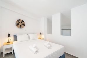 Quarto de hotel com cama, duas mesinhas de cabeceira, uma luminária fixada na parede, uma peça decorativa circular feita de fibras naturais e uma janela.