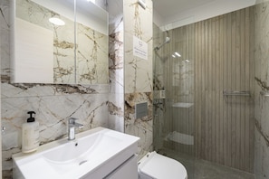 Casa de banho com painel de parede de mármore, lavatório branco, sanita e cabine de duche com parede de vidro.