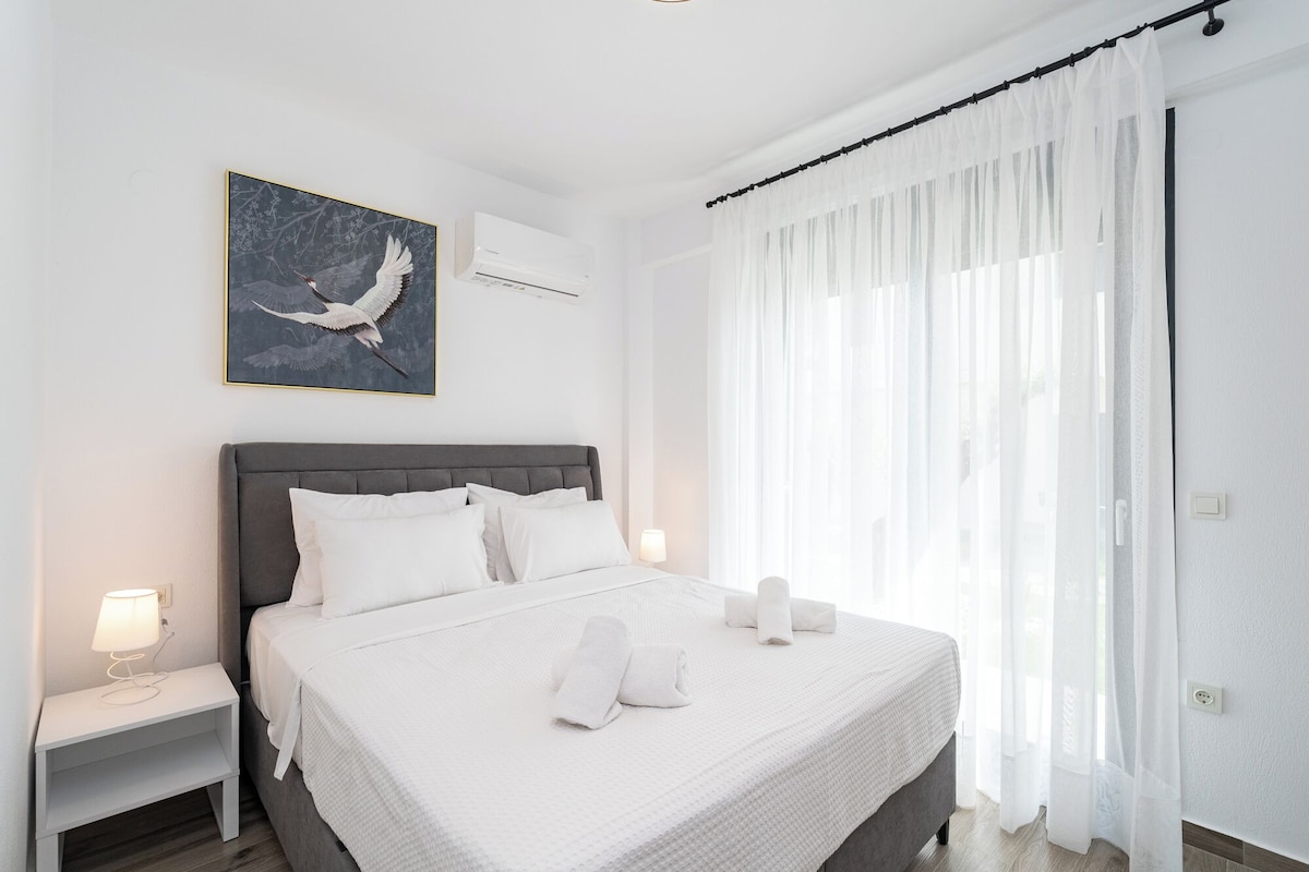 Quarto de hotel com cama, roupa de cama branca, mesa de cabeceira com abajur, pintura de pássaros e cortinas finas.