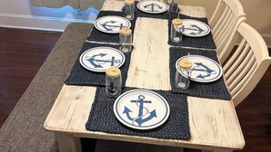 A dining table with nautical-themed placemats and salt shakers, surrounded by white chairs. - Charming Galveston home near cruise terminal, beach, UTMB & downtown attractions (Galveston)