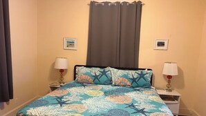 A bed with a patterned bedspread, two bedside tables with lamps, a window with curtains, and framed pictures on the wall. - Charming Galveston home near cruise terminal, beach, UTMB & downtown attractions (Galveston)