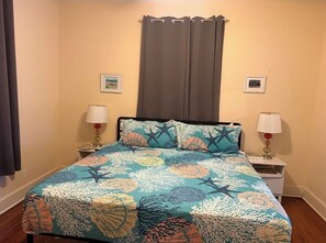 A bed with a patterned bedspread, two bedside tables with lamps, a window with curtains, and framed pictures on the wall. - Charming Galveston home near cruise terminal, beach, UTMB & downtown attractions (Galveston)
