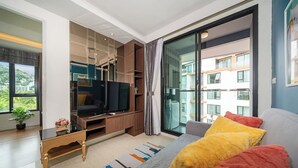 A modern living room with a flat-screen TV, a sofa with colorful cushions, a coffee table, and a large sliding glass door leading to a balcony. - Best for Digital Nomad-1 BR Apt close to Surin Beach (Tambon Choeng Thale)