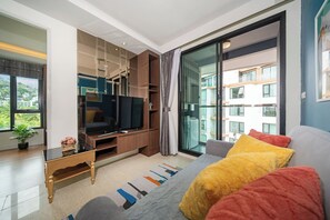 Ein modernes Wohnzimmer mit einem Flachbildfernseher, einer Couch mit bunten Kissen, einem Couchtisch und einer großen Schiebeglastür, die auf einen Balkon führt.