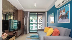 A modern living room with a flat-screen TV, a sofa with colorful cushions, and a large window with a view of buildings. - Best for Digital Nomad-1 BR Apt close to Surin Beach (Tambon Choeng Thale)