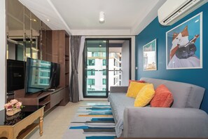 Ein modernes Wohnzimmer mit einem Flachbildfernseher, einer Couch mit bunten Kissen und einem großen Fenster mit Blick auf Gebäude.