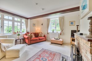 Ein Wohnzimmer mit einem roten Sofa, einem weißen Sessel, einem gemusterten Teppich, einem Kamin und großen Fenstern mit Vorhängen.
