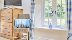 A room with a wooden dresser, a wicker chair, a window with curtains, and a television mounted on the wall. - Piddlehinton Coach House - Piddlehinton, Dorchester, sleeps 6 guests (Piddlehinton)