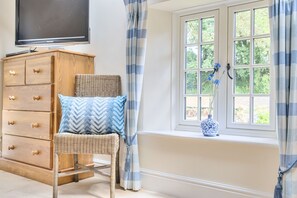 A room with a wooden dresser, a wicker chair, a window with curtains, and a television mounted on the wall. - Piddlehinton Coach House - Piddlehinton, Dorchester, sleeps 6 guests (Piddlehinton)