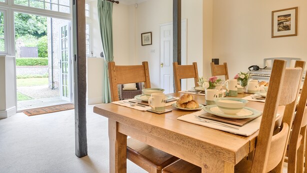 A dining room with a wooden table set for breakfast, chairs, and a view of the outdoors through a door. - Piddlehinton Coach House - Piddlehinton, Dorchester, sleeps 6 guests (Piddlehinton)