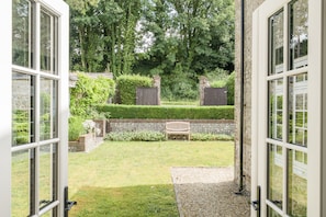 Eine geöffnete Tür, die in einen gepflegten Garten mit Bank, Hecke und Backsteinmauer führt.