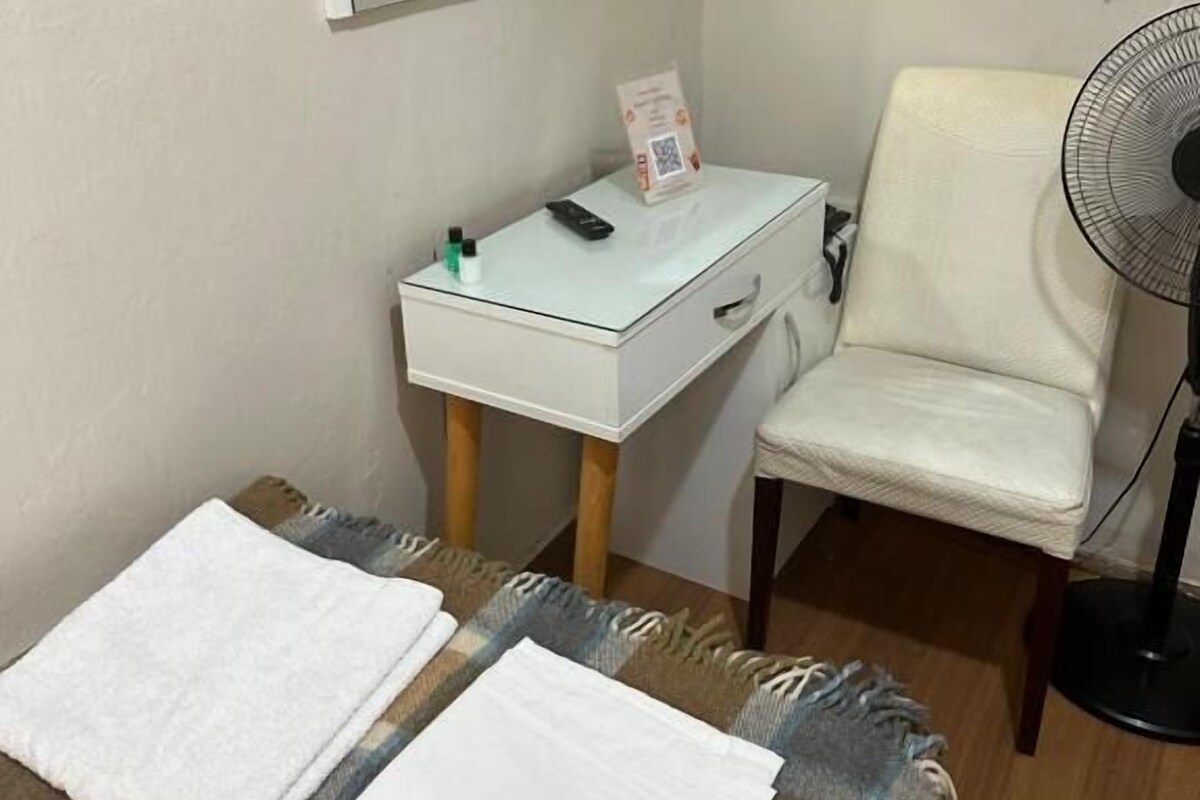 A small room with a bed, a bedside table with a remote control, a chair, a standing fan, and a television mounted on the wall.