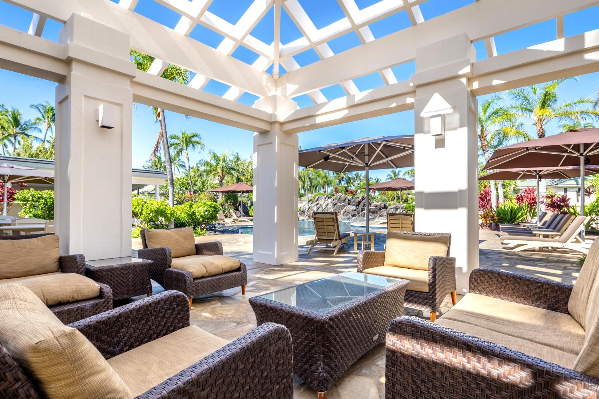 A covered outdoor lounge area with wicker furniture, a glass-top table, and umbrellas providing shade.