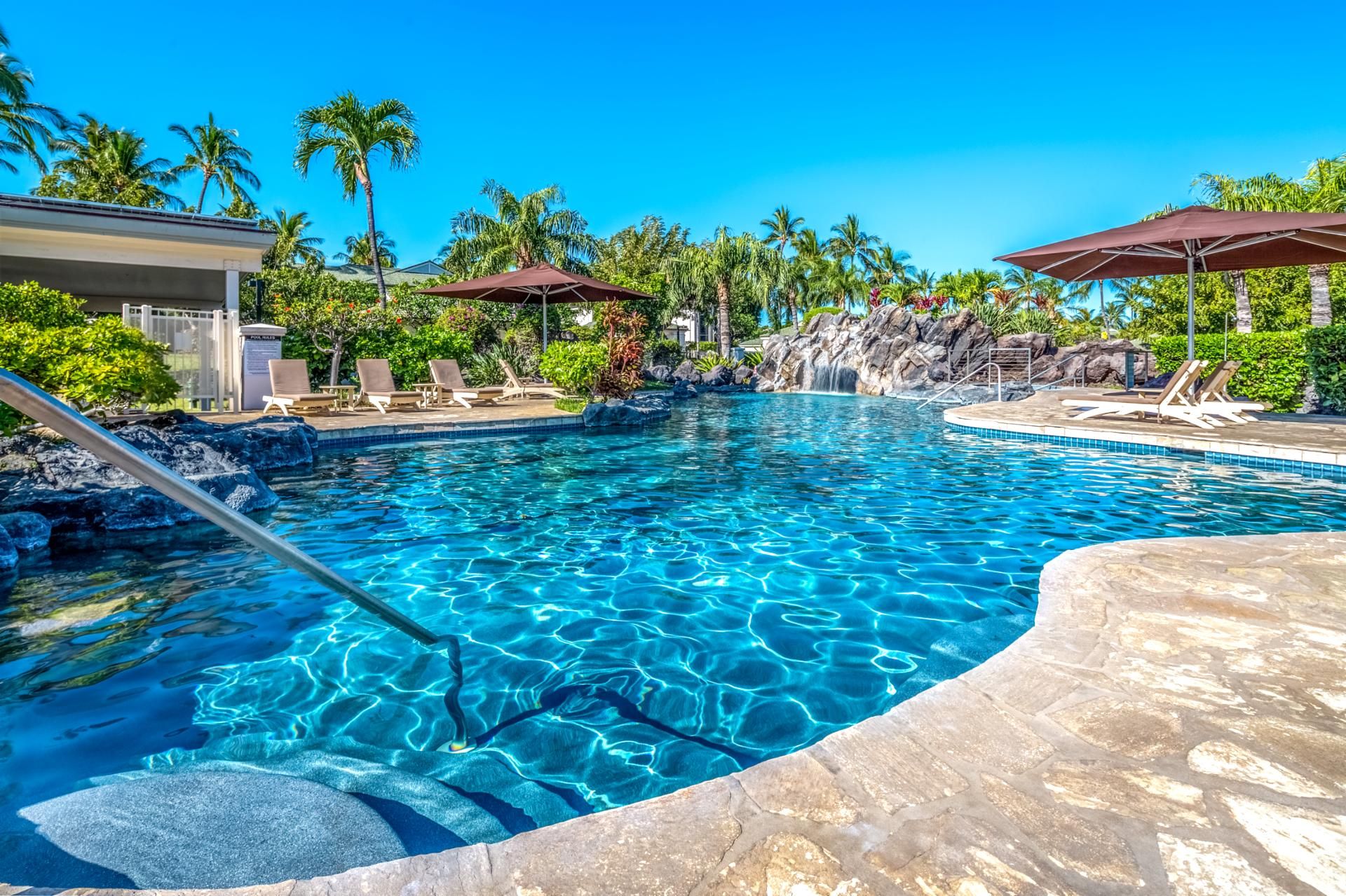 A clear blue pool with lounge chairs and umbrellas, surrounded by palm trees and a waterfall feature.
