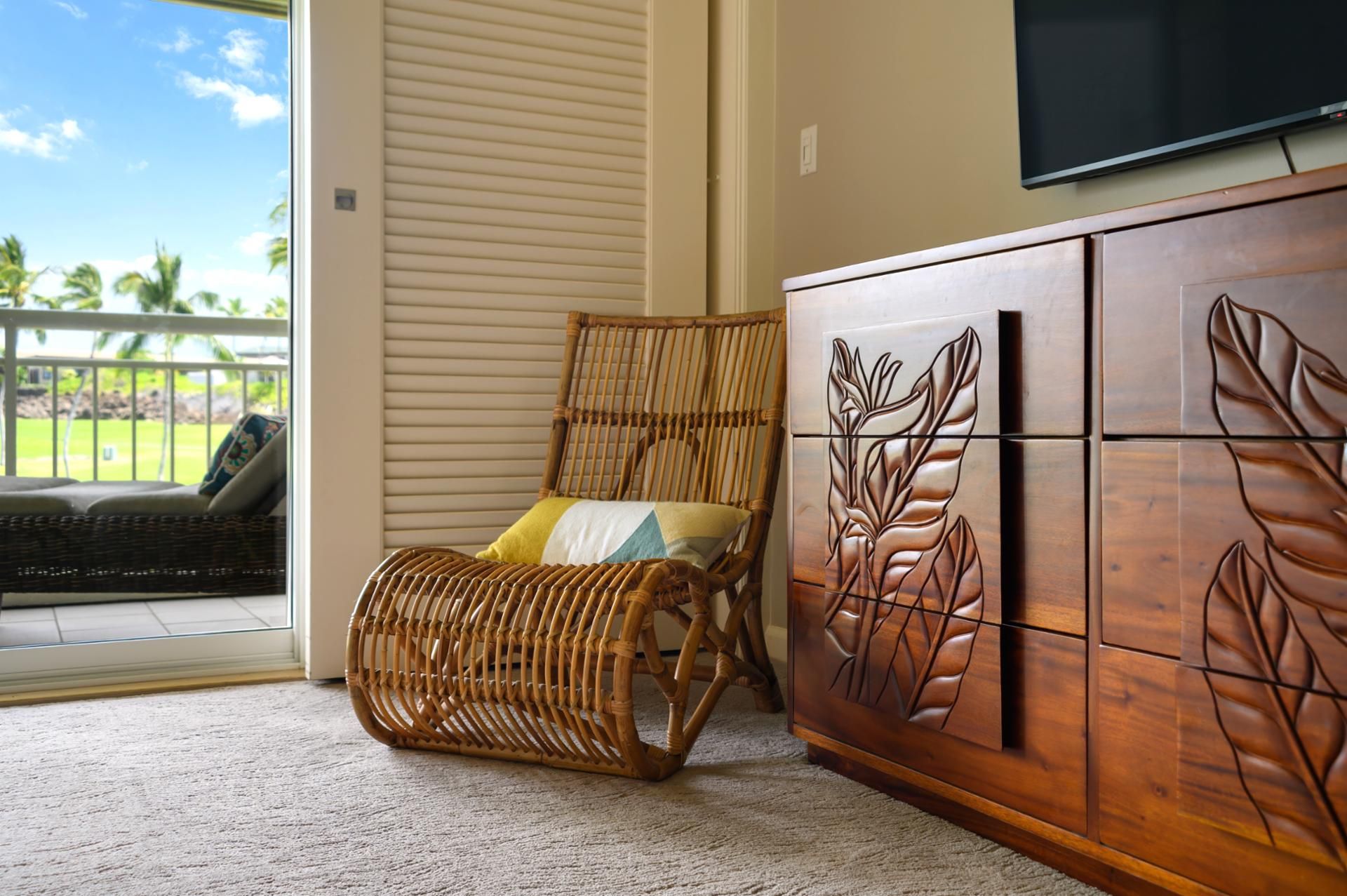 A wicker chair with cushions, a wooden cabinet with leaf designs, and a television mounted on the wall.