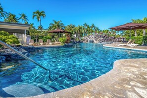 Uma piscina azul límpida com espreguiçadeiras e guarda-sóis, cercada por palmeiras e uma paisagem rochosa.