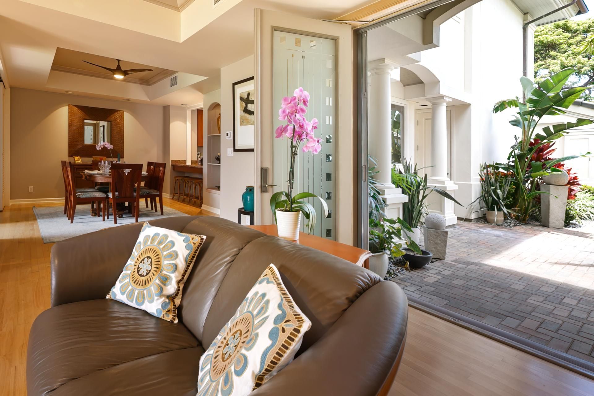 A modern living room with a brown leather sofa, a dining area with wooden furniture, and a view of a garden with various plants.