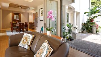 A modern living room with a brown leather sofa, a dining area with wooden furniture, and a view of a garden with various plants.