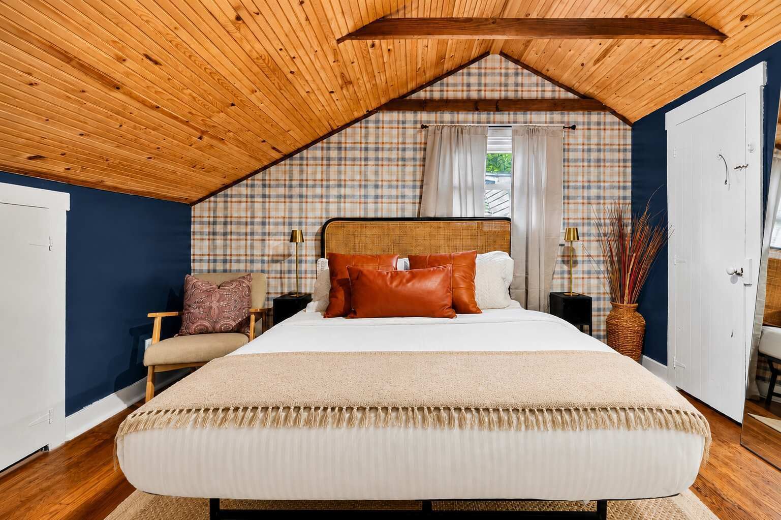 Quarto com teto de madeira, uma cama grande, mesa de cabeceira, uma cadeira e uma janela com cortinas.