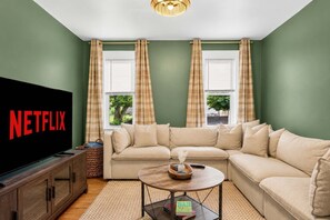 Ein Wohnzimmer mit einem Ecksofa, einem Fernseher mit Netflix-Logo und zwei mit Vorhängen versehenen Fenstern.