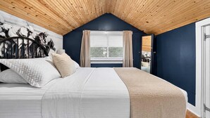 Una camera da letto con soffitto spiovente, un letto con biancheria bianca, una finestra con tende e vista sull'esterno.
