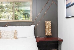 Ein Bett mit weißer Bettwäsche, eine gewebte Lampe und ein Fenster mit Jalousien.
